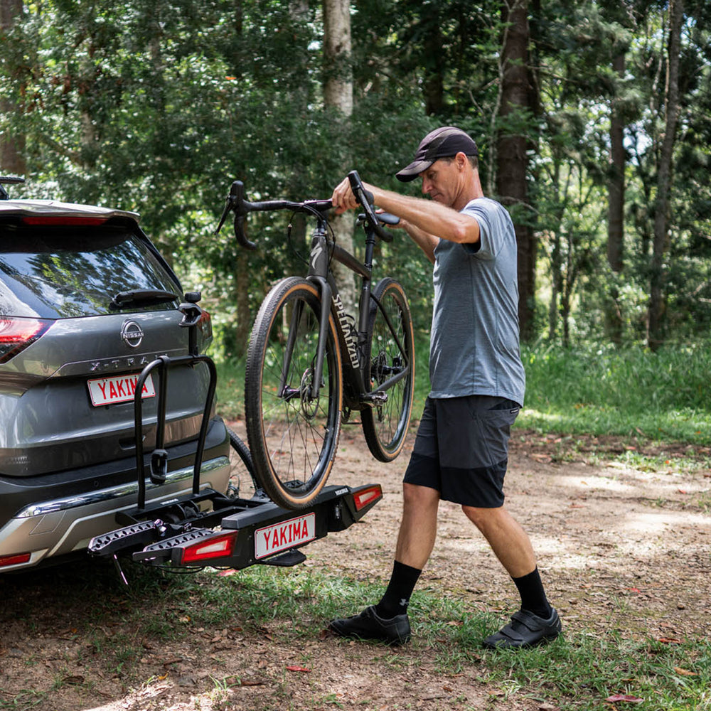 Man loading a bicycle onto a car with a Yakima bike rack in a forest setting.